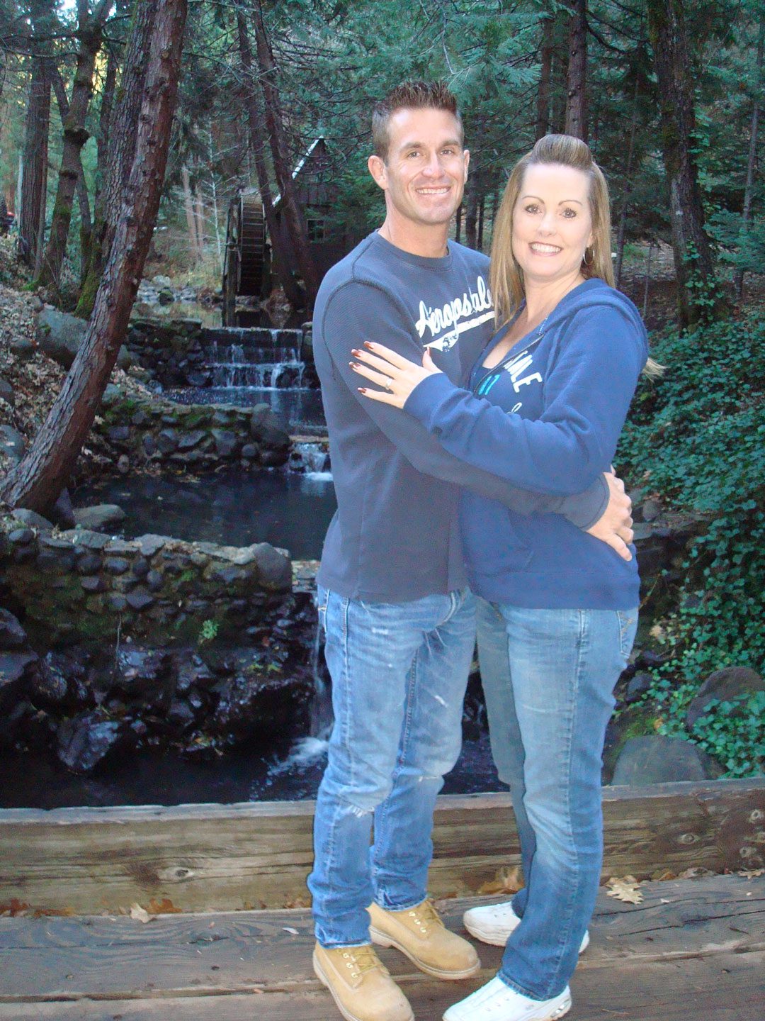 A man and a woman standing close together and smiling in front of a small waterfall and wooden structure, surrounded by trees and greenery.